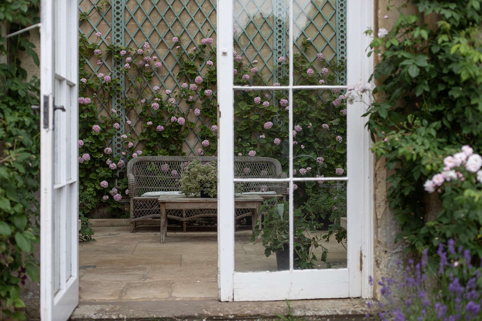 An inviting garden seating area surrounded by climbing roses and greenery, visible through white open French doors.