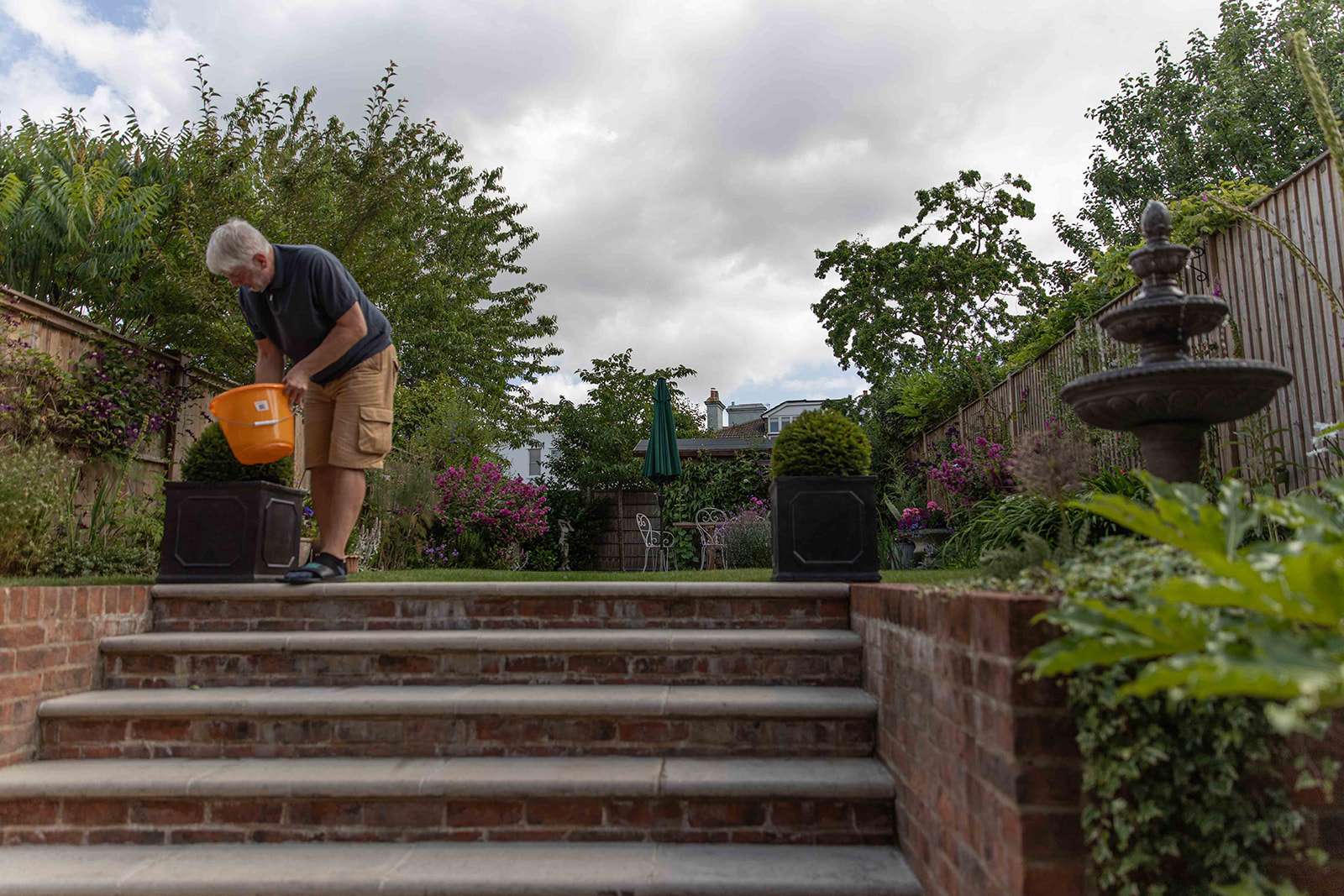 A person watering plants on a brick stairway leading to a lush garden with a fountain and outdoor furniture in the background.