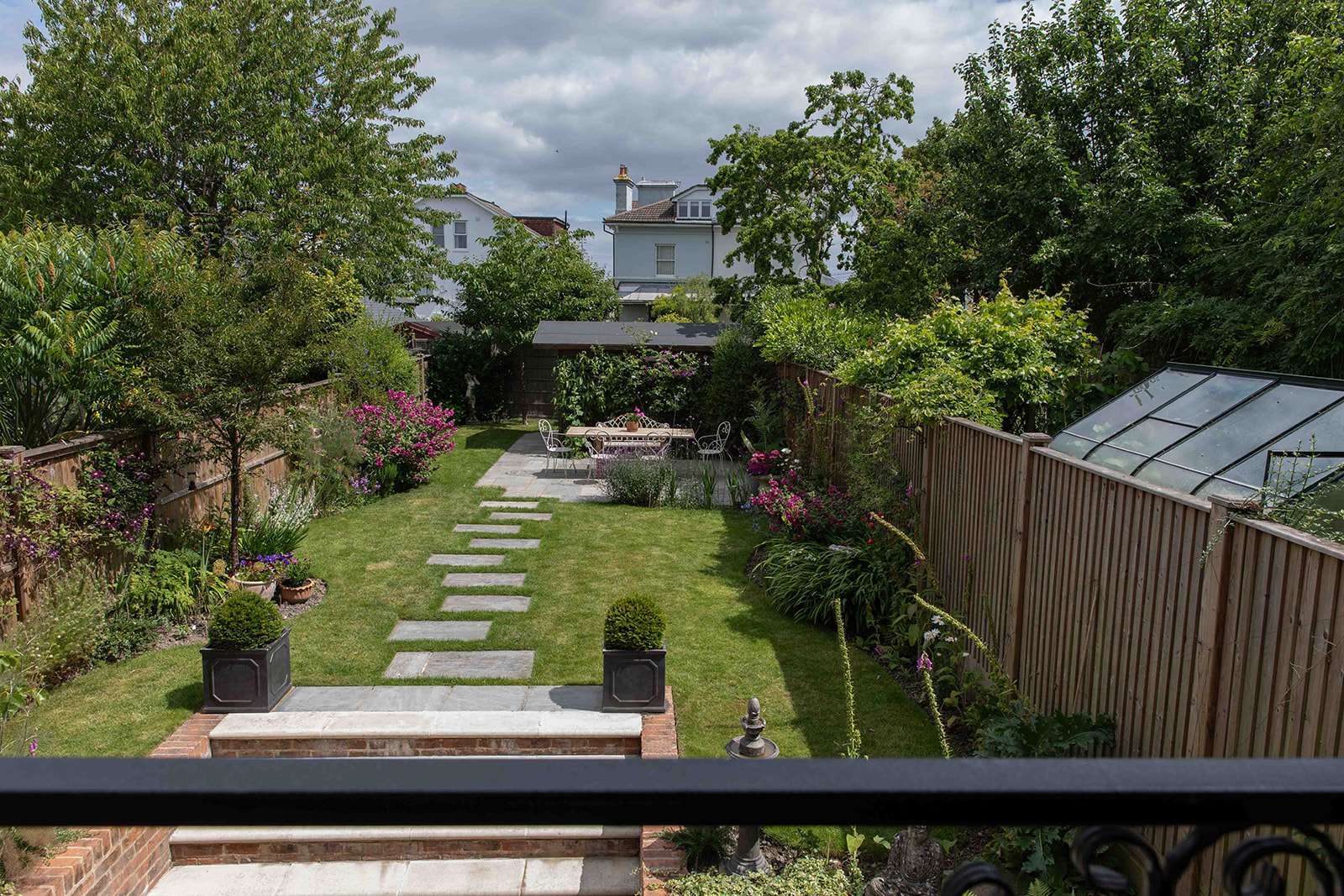 Lush backyard garden with stepping stones leading to a seating area, surrounded by flowering shrubs and a greenhouse under a cloudy sky
