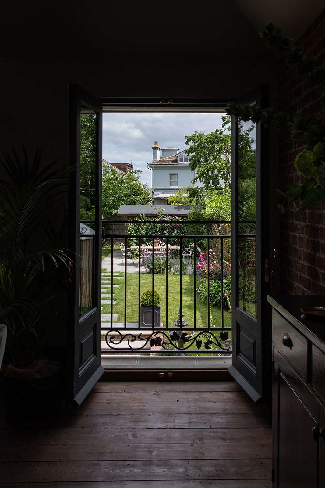 View from inside a house showing an open door leading to a lush garden with a wrought-iron gate, surrounded by greenery and a house in the background.