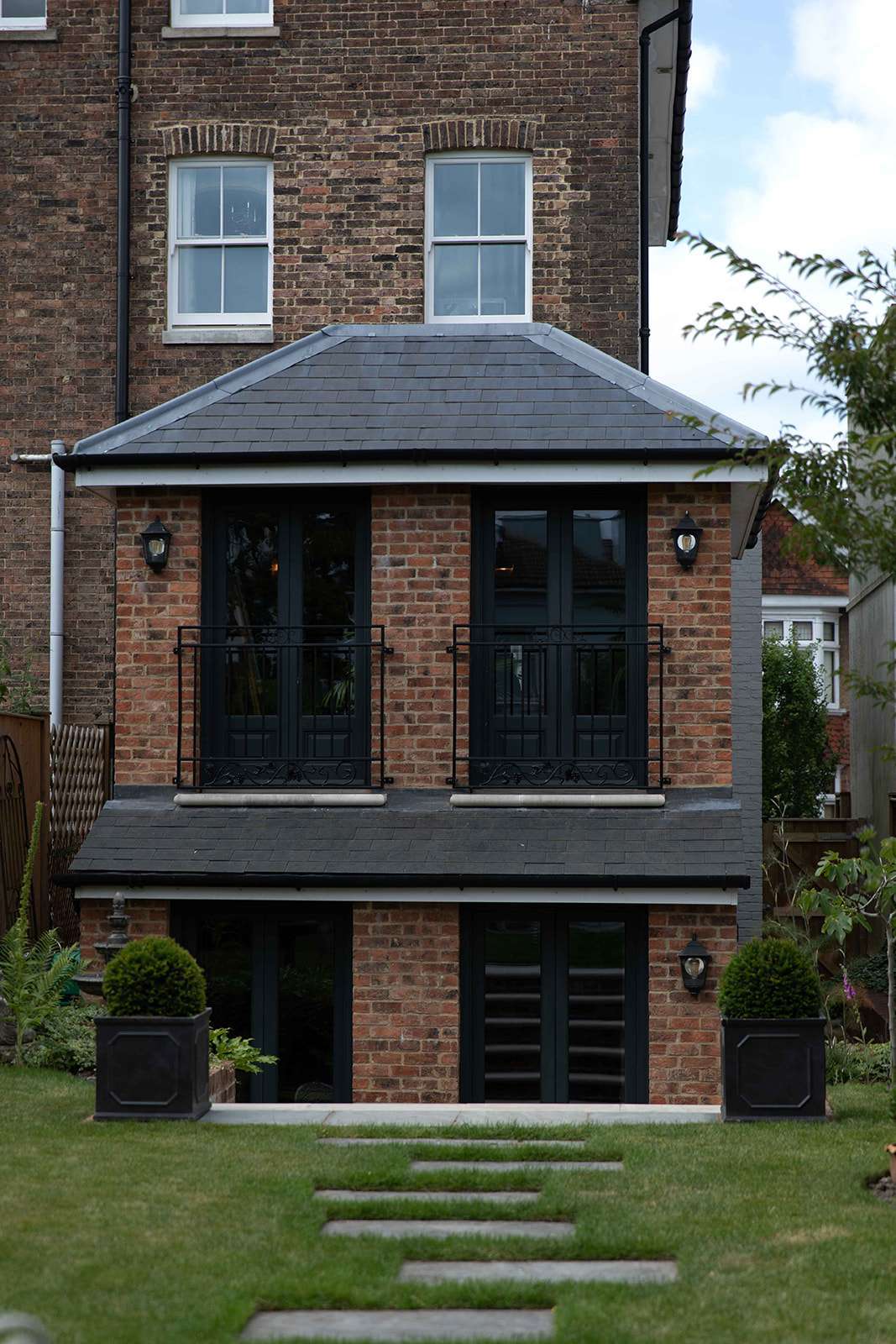 A two-story brick house with a modern extension featuring large windows and a balcony with a metal railing, set in a landscaped garden with stepping stones.