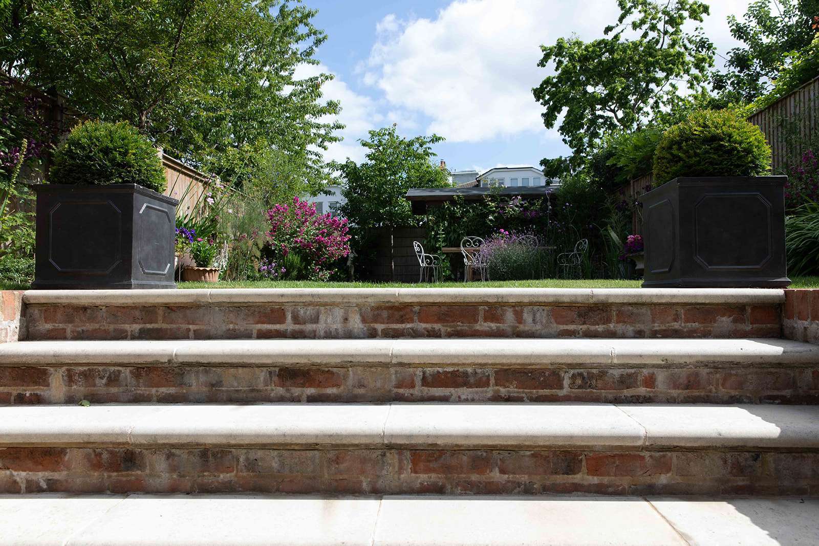 A well-manicured garden with a flight of brick and stone steps flanked by large planters and blooming flowers in the background.
