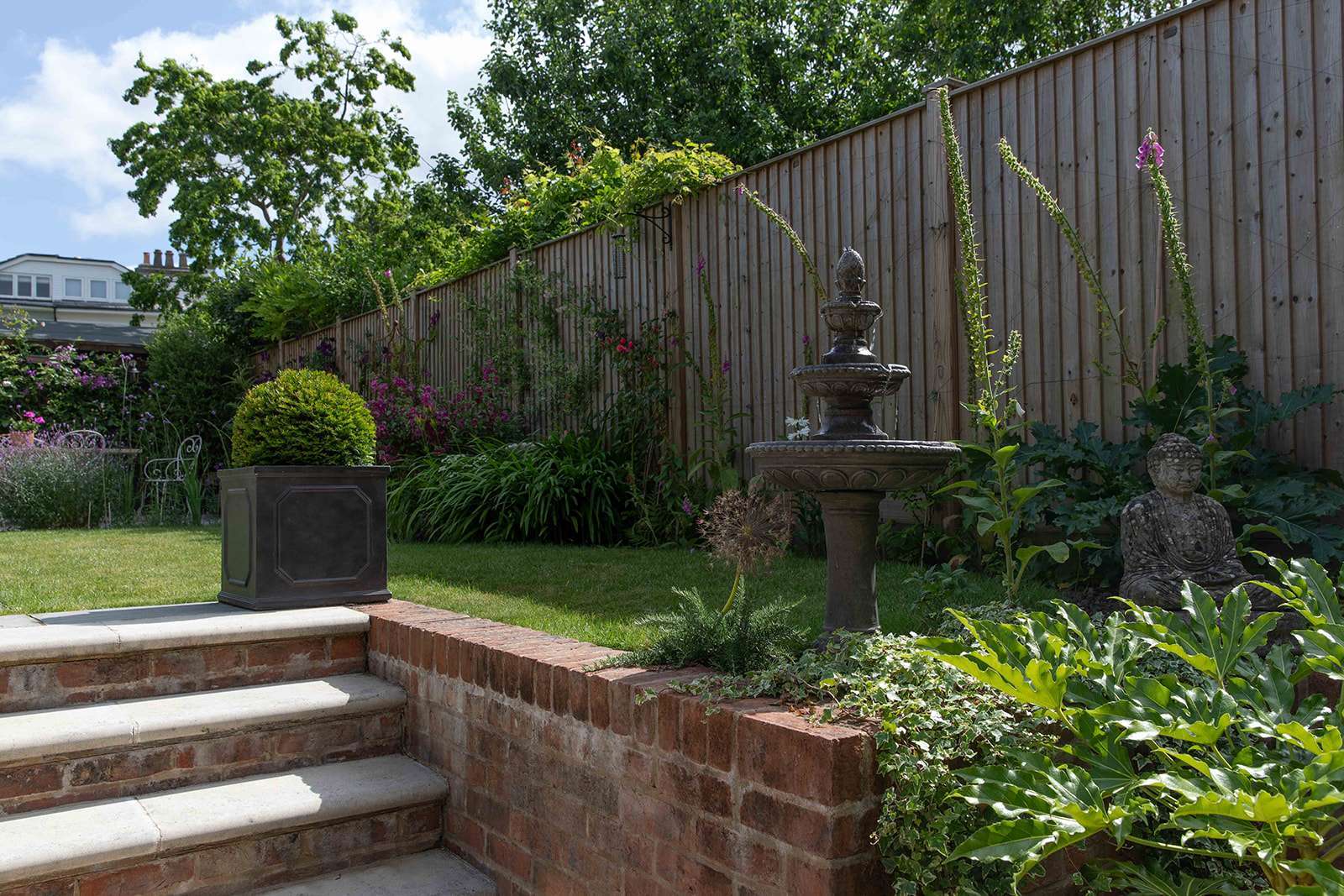 A serene garden with a stone fountain, Buddha statue, vibrant plants, and a wooden fence in the background.