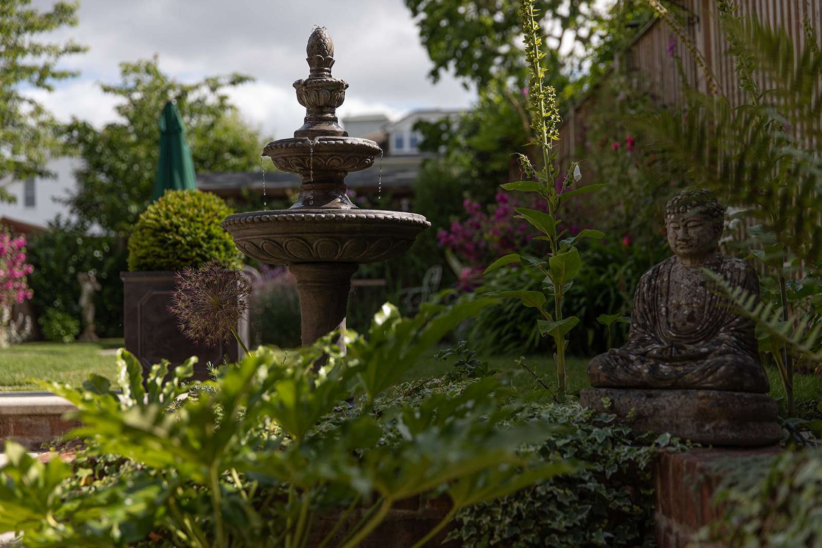 A tranquil garden scene featuring a multi-tiered fountain in the background with water gently cascading down, and a weathered Buddha statue sitting peacefully among the lush greenery in the foreground.