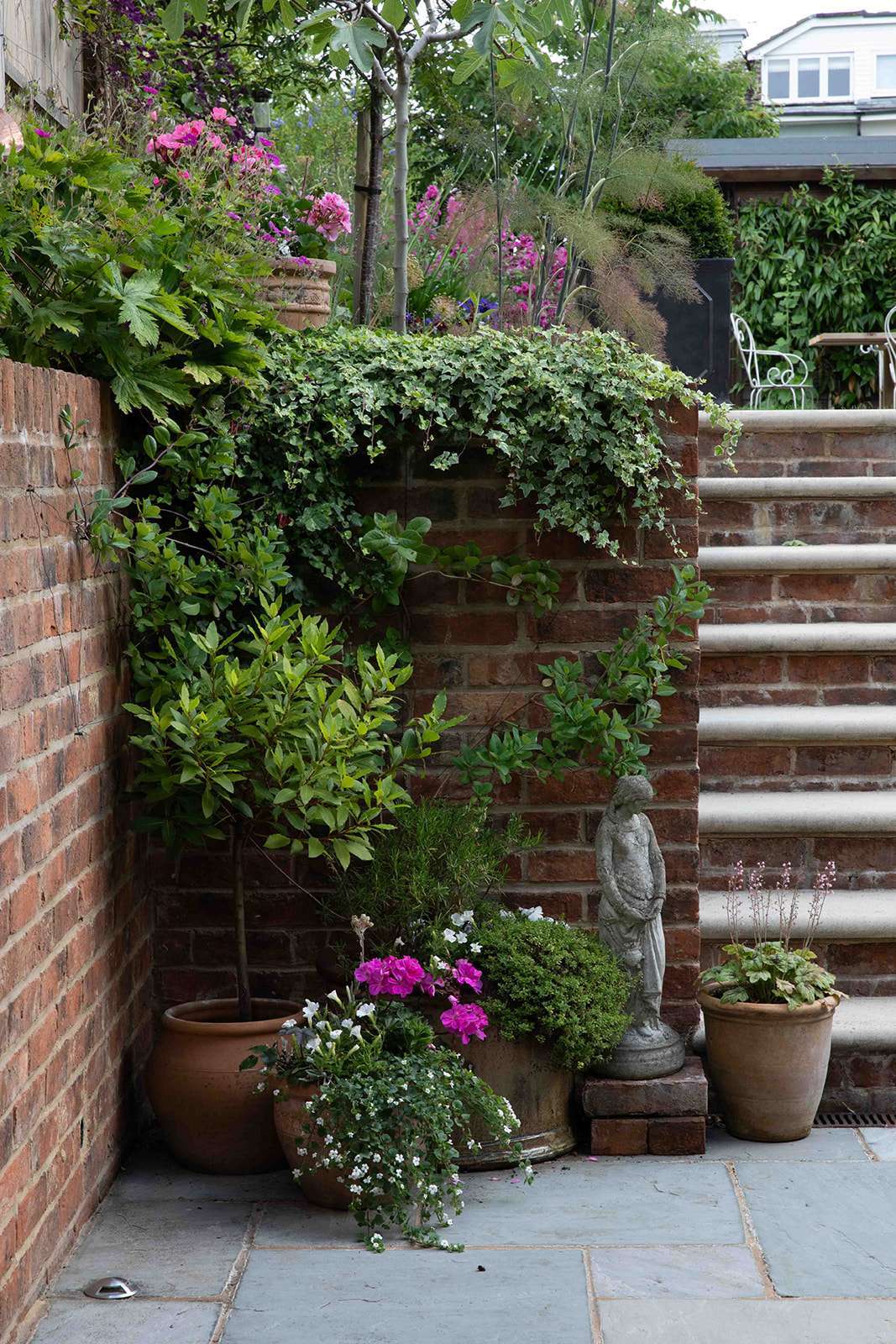 A serene corner in a garden showcasing an assortment of plants, a brick wall covered in creeping ivy, terracotta pots with blooming flowers, and a stone statue, all next to a set of stone steps.