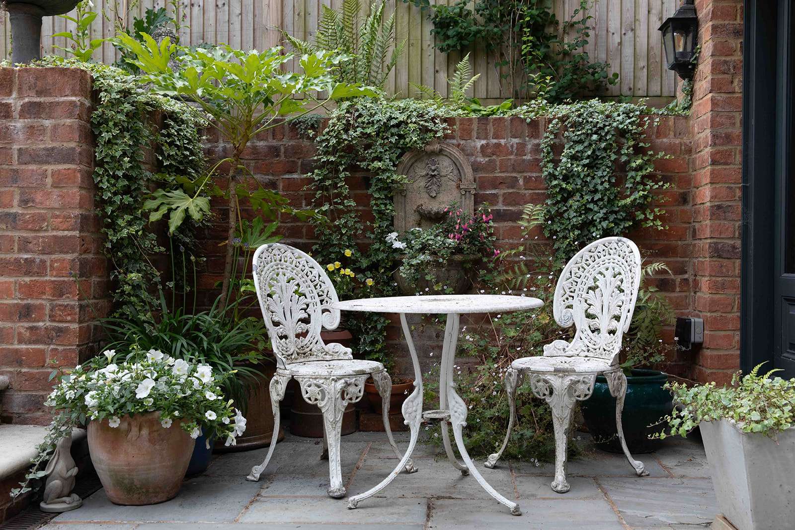An intimate brick patio with vintage white metal table and chairs, surrounded by lush greenery and ivy-covered walls.