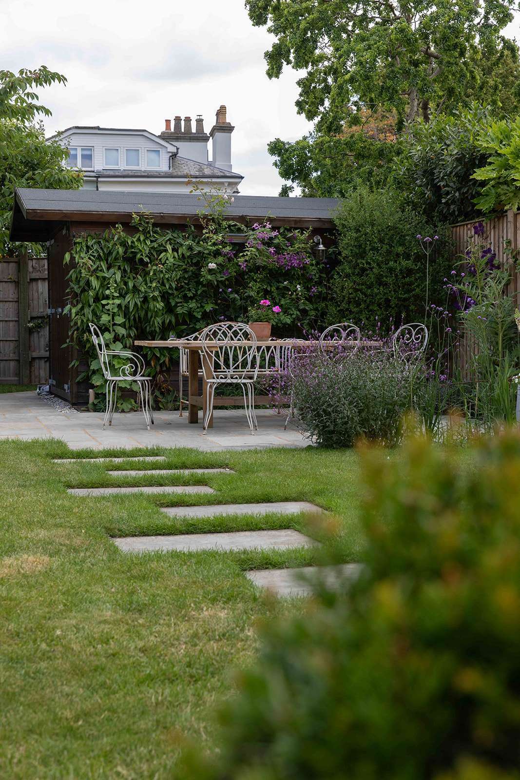 A peaceful garden setting with a white wrought iron table and chairs, stepping stones on the grass leading to a patio, and surrounding greenery and flowers, with a house partially visible in the background.