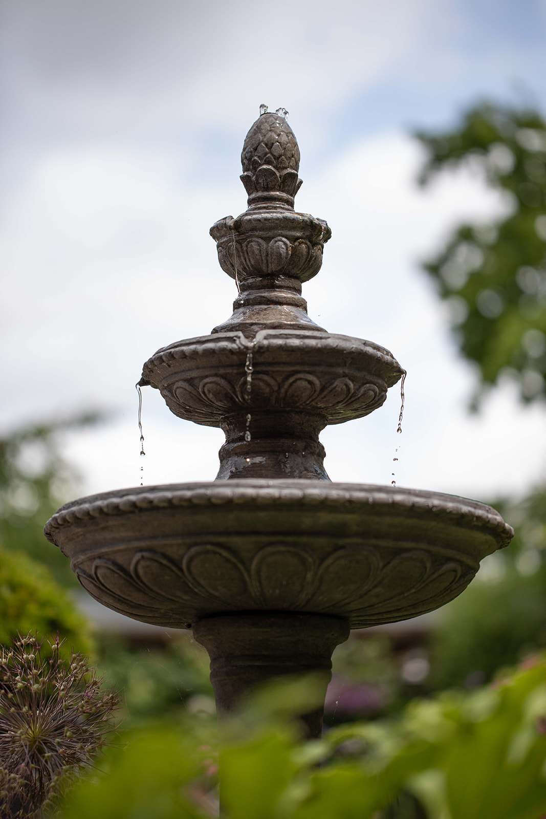Close-up of an ornate garden fountain with water gently dripping from its tiered structure, surrounded by greenery.