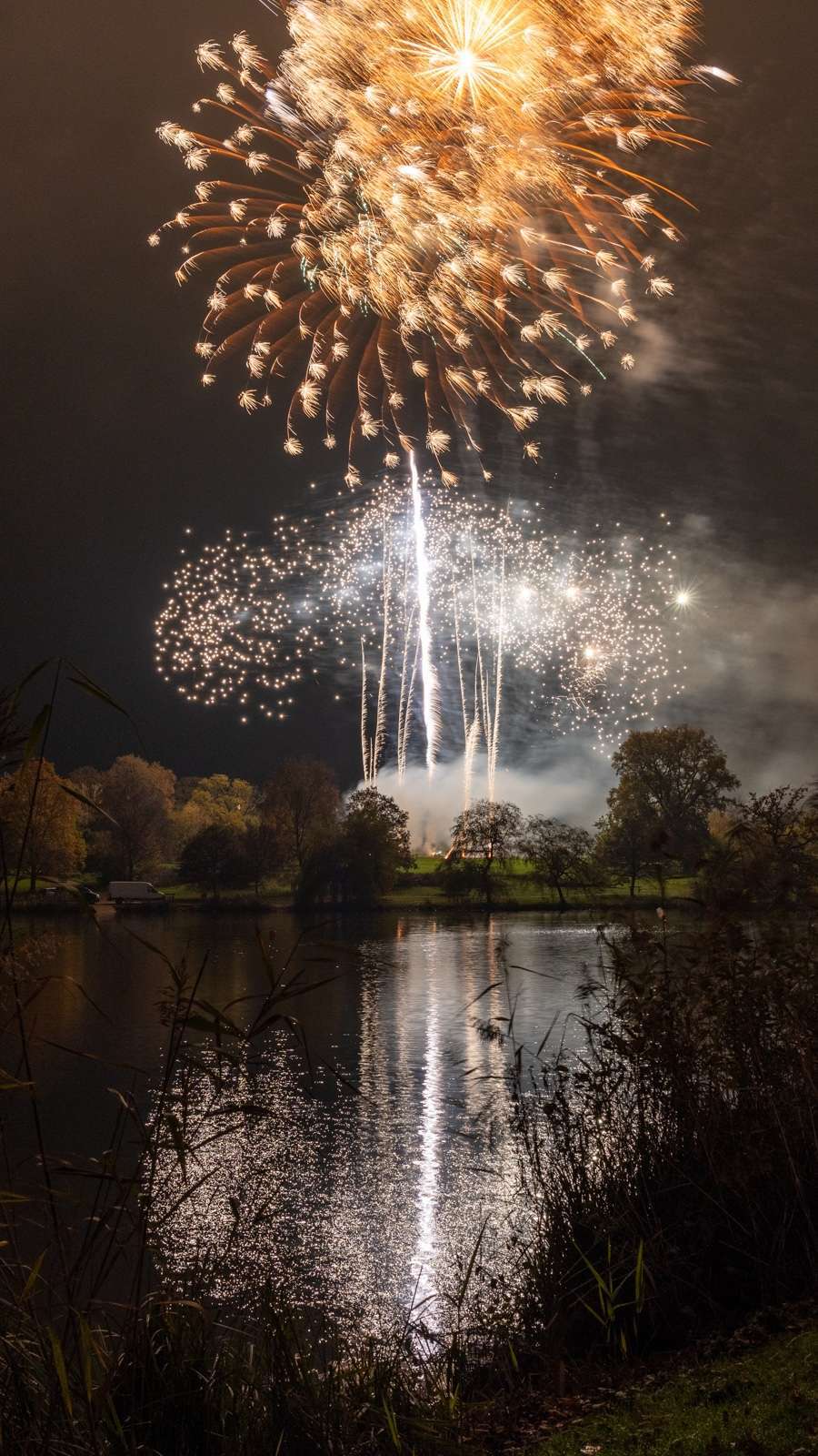 Fireworks display illuminates the night sky above a lake with reflections on the water surface, framed by dark foliage.